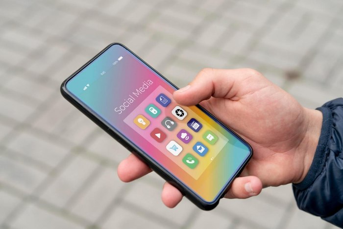 Roka drži sodoben pametni telefon, na katerem je viden barvit zaslon z naloženimi mobilnimi aplikacijami.